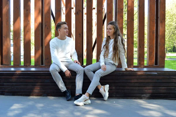 A young girl and a young man in the same clothes sitting on a wooden bench. — Stock Photo, Image