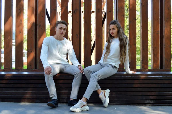 A young girl and a young man in the same clothes sitting on a wooden bench. — Stock Photo, Image