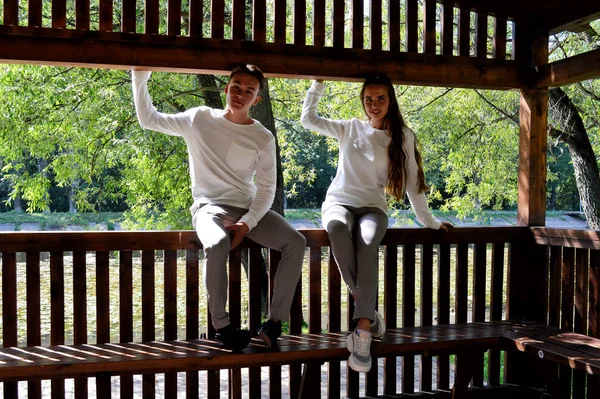 Uma jovem e um jovem com as mesmas roupas sentados em um banco de madeira . — Fotografia de Stock