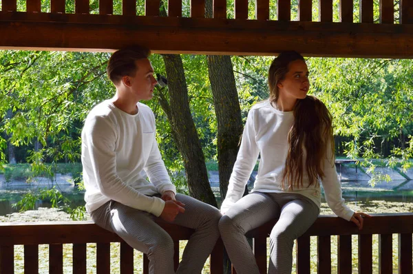 En ung flicka och en ung man i samma kläder sitter på en träbänk. — Stockfoto