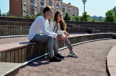 Genç bir kız ve aynı kıyafetleri giyen genç bir adam ahşap bir bankta oturuyor.