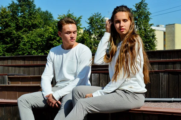 Genç bir kız ve aynı kıyafetleri giyen genç bir adam ahşap bir bankta oturuyor.