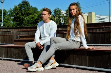 Genç bir kız ve aynı kıyafetleri giyen genç bir adam ahşap bir bankta oturuyor.