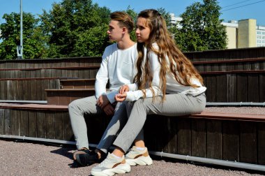 Genç bir kız ve aynı kıyafetleri giyen genç bir adam ahşap bir bankta oturuyor.