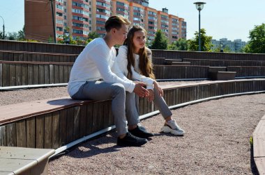 Genç bir kız ve aynı kıyafetleri giyen genç bir adam ahşap bir bankta oturuyor.