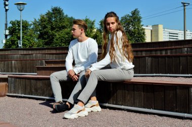 Genç bir kız ve aynı kıyafetleri giyen genç bir adam ahşap bir bankta oturuyor.