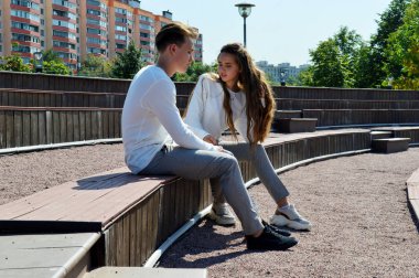 Genç bir kız ve aynı kıyafetleri giyen genç bir adam ahşap bir bankta oturuyor.