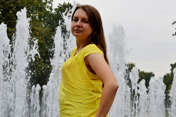 一个黑头发的女人穿着黄色的T恤站在喷泉边 — 图库照片