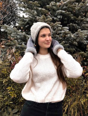 Siyah saçlı, beyaz kazaklı, gri şapkalı ve gri eldivenli genç bir kız kışın sokakta bir Noel ağacının arka planında