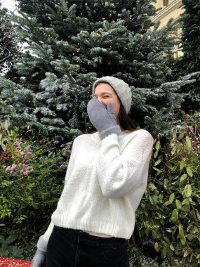 Siyah saçlı, beyaz kazaklı, gri şapkalı ve gri eldivenli genç bir kız kışın sokakta bir Noel ağacının arka planında