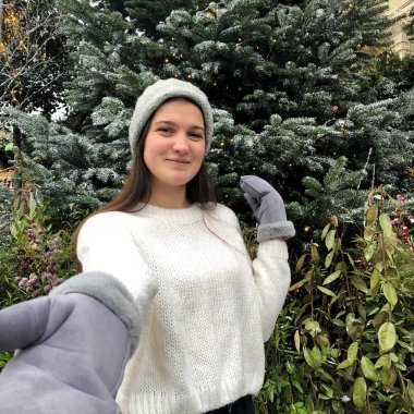 Siyah saçlı, beyaz kazaklı, gri şapkalı ve gri eldivenli genç bir kız kışın sokakta bir Noel ağacının arka planında