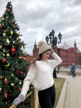 Siyah saçlı, beyaz kazaklı, bej şapkalı ve gri eldivenli genç bir kız kışın sokakta bir Noel ağacının arka planında