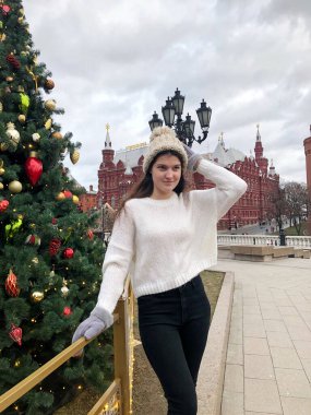 Siyah saçlı, beyaz kazaklı, bej şapkalı ve gri eldivenli genç bir kız kışın sokakta bir Noel ağacının arka planında