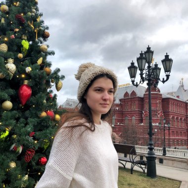 Siyah saçlı, beyaz kazaklı, bej şapkalı ve gri eldivenli genç bir kız kışın sokakta bir Noel ağacının arka planında