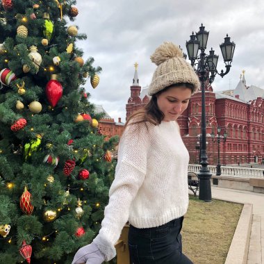 Siyah saçlı, beyaz kazaklı, bej şapkalı ve gri eldivenli genç bir kız kışın sokakta bir Noel ağacının arka planında