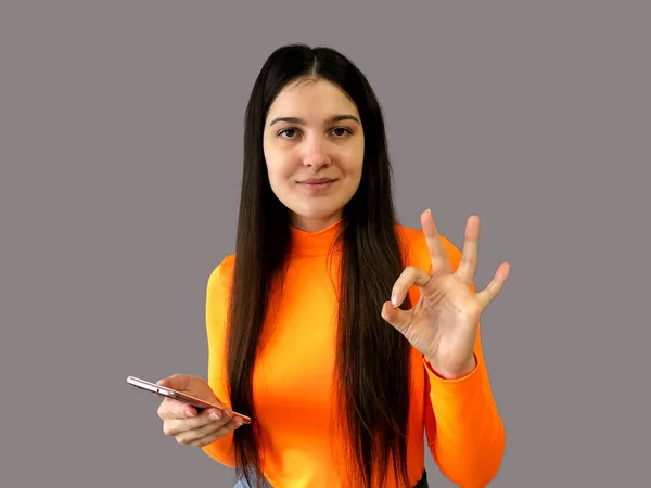 Młoda Piękna Dziewczyna Ciemnymi Włosami Jasnopomarańczowym Swetrze Szarym Tle Telefonem — Zdjęcie stockowe