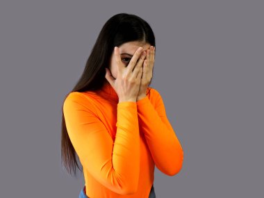 Gri arka planda parlak turuncu süveter giyen koyu renk saçlı güzel bir kız. Yüzünü kendi elleriyle kapatıyor.