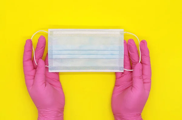 Disposable latex glove. The pink gloves. The nurse\'s gloved hands are on the table. Disposable surgical blue mask on the table. Security during the quarantine period. Yellow background.