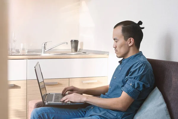 一位长相像亚洲人的年轻人穿着蓝色衬衫 牛仔裤 黑头发 在家里笔记本电脑后面工作 办公室外的远程工作 商业与教育 — 图库照片