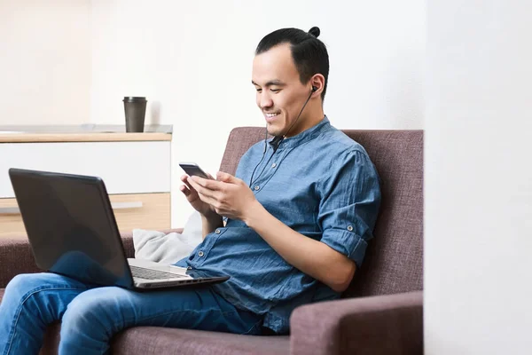 一个亚洲人看着电话笑着 白天在家里沙发上的耳机里和笔记本电脑一起工作 办公室外的远程工作 业余爱好和休闲 — 图库照片