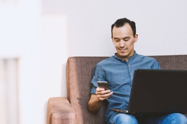 Genç bir adam telefona ve laptopa bakıyor. Evde ve iş yerinde çalış. İletişim ve teknoloji