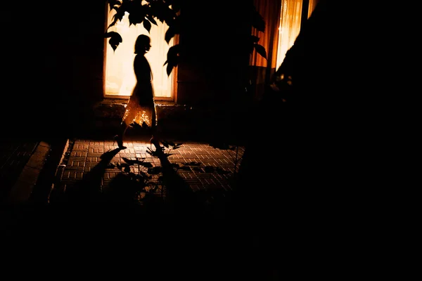 Silhouette of a young girl on a background of a bright window in the evening. In the foreground foliage and trees