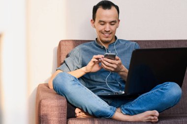 Genç yakışıklı bir Asyalı işadamı evde kanepede otururken gülümsüyor ve bir telefon tutuyor. Yakın arkadaşlarla çevrimiçi çalışmak ve iletişim kurmak