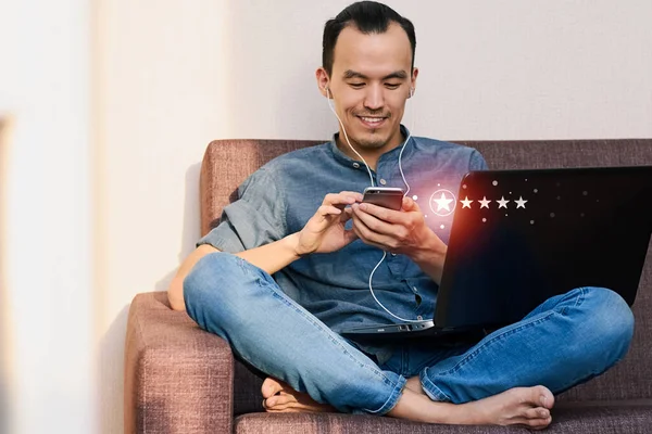 一位面带微笑的年轻亚洲男子手持装有五星级反馈评级图标的智能手机 这个家伙坐在家里的沙发上 对商务服务的概念给出了很好的评价 — 图库照片