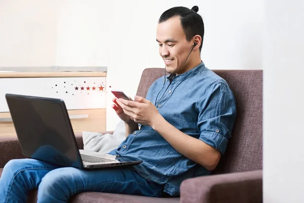 一位面带微笑的年轻亚洲男子手持装有五星级反馈评级图标的智能手机 这家伙坐在家里的沙发上 对商务服务的概念给予了很高的评价 — 图库照片