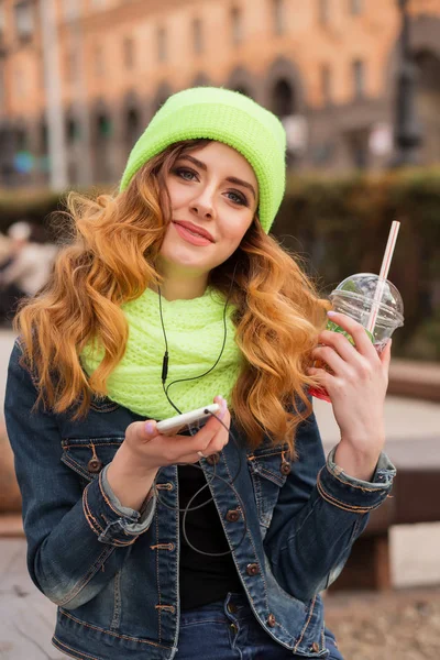 Mujer joven feliz con un teléfono móvil en la mano, gorra verde claro y bufanda, beber cóctel al aire libre —  Fotos de Stock