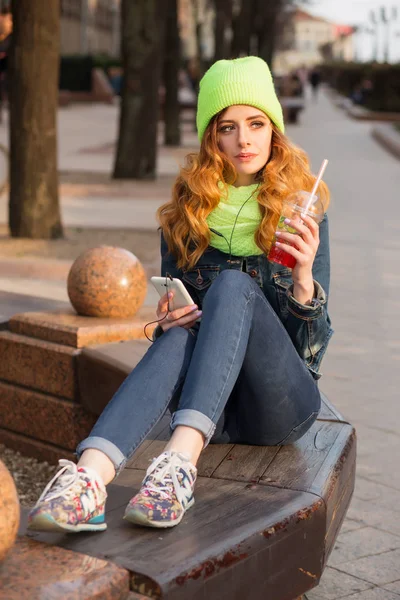Mujer joven feliz con un teléfono móvil en la mano, gorra verde claro y bufanda, beber cóctel al aire libre —  Fotos de Stock