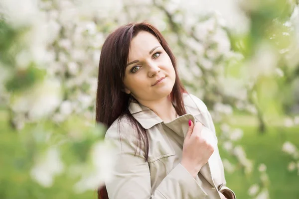 Schöne junge brünette Frau, die an einem warmen Frühlingstag neben dem blühenden Apfelbaum steht — Stockfoto