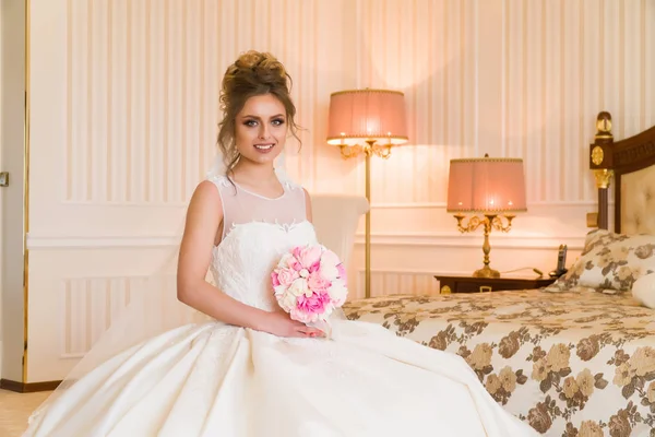 Portret van mooie jonge bruid. Een meisje is poseren in een hotelkamer. Een dame zit met een boeket rozen bij het raam. bruid wachten in hotel — Stockfoto