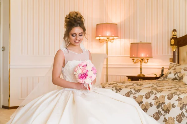 Portret van mooie jonge bruid. Een meisje is poseren in een hotelkamer. Een dame zit met een boeket rozen bij het raam. bruid wachten in hotel — Stockfoto