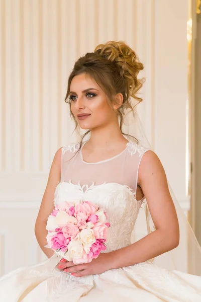 Portret van mooie jonge bruid. Een meisje is poseren in een hotelkamer. Een dame zit met een boeket rozen bij het raam. bruid wachten in hotel — Stockfoto