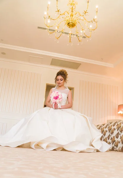 Portret van mooie jonge bruid. Een meisje is poseren in een hotelkamer. Een dame zit met een boeket rozen bij het raam. bruid wachten in hotel — Stockfoto