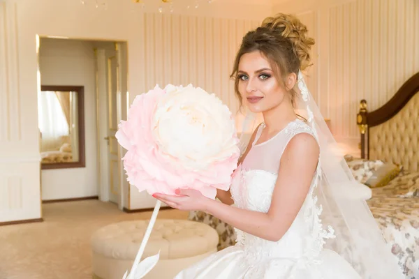 Portret van mooie jonge bruid. Een meisje is poseren in een hotelkamer. Een dame zit met een papieren bloem bij het raam. bruid wachten in hotel — Stockfoto