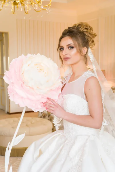 Portret van mooie jonge bruid. Een meisje is poseren in een hotelkamer. Een dame zit met een papieren bloem bij het raam. bruid wachten in hotel — Stockfoto