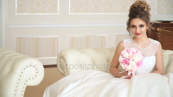 Привлекательная молодая женщина невеста с букетом цветов сидит на диване в роскошном номере. Она ждет кого-то с волнением — стоковое видео