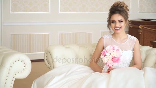 Μια ελκυστική νεαρή γυναίκα νύφη με ένα μπουκέτο από λουλούδια είναι κάθεται στον καναπέ στο πολυτελές δωμάτιο. Είναι σε αναμονή για κάποιον με ενθουσιασμό — Αρχείο Βίντεο