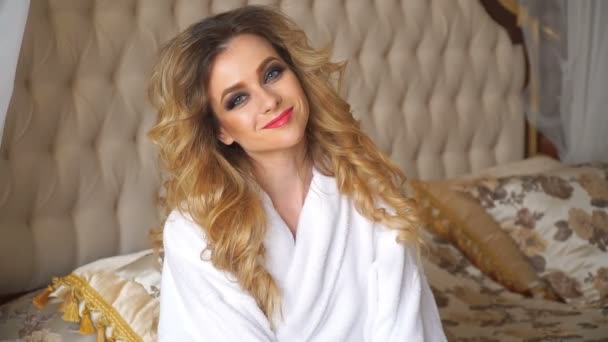 Schöne verführerische Frau flirtet mit der Kamera, sitzt auf einem Bett im weißen Bademantel und blickt kokett nach oben — Stockvideo