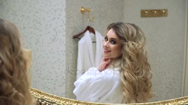Güzel sarışın kız saçını ve onun banyo aynaya bakarak düzeltir. Güzellik genç kadın saçlarını düzeltir. Saç şekillendirme