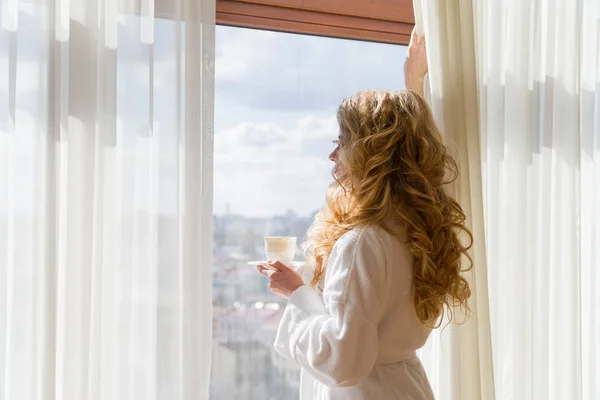 Piękna dziewczyna picia kawy. Piękna kobieta, otwierając zasłony, patrząc przez okno i ciesząc się poranną kawę — Zdjęcie stockowe