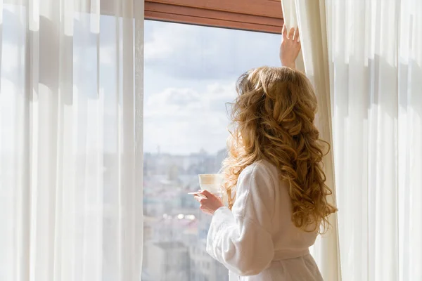 Beauty girl drinking coffee. Beautiful woman opening curtains, looking out the window and enjoying her morning coffee