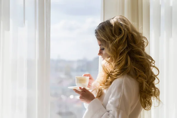 Belle fille buvant du café. Belle femme ouvrant les rideaux, regardant par la fenêtre et profitant de son café du matin — Photo