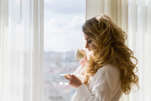 Schönheit Mädchen Kaffee trinken. schöne Frau öffnet Vorhänge, schaut aus dem Fenster und genießt ihren Morgenkaffee — Stockfoto