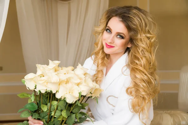 Belle femme séduisante flirtant avec la caméra est assise sur le lit avec un grand bouquet de roses blanches en peignoir blanc levant les yeux avec un look coquette — Photo