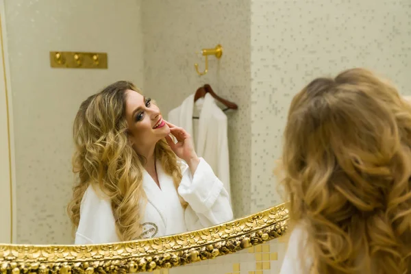 Hermosa chica rubia corrige su cabello y se mira en el espejo en su baño. Belleza mujer joven corrige su cabello. Peinado — Foto de Stock