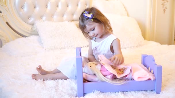 An active little preschool child, a pretty little girl with a blond curly hair, plays with her dolls, puts them to sleep in — Stock Video