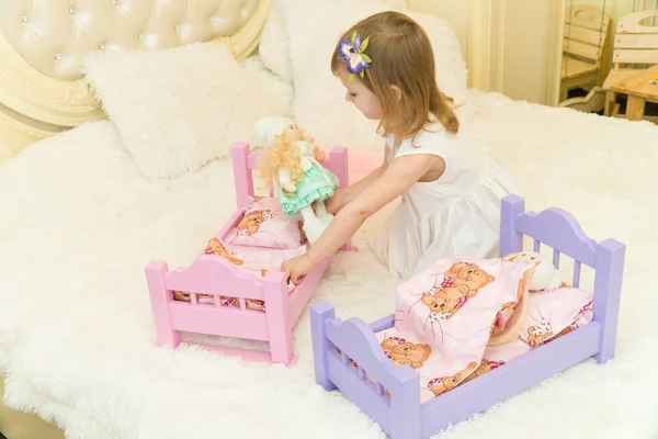 Uma criança pré-escolar ativa, uma menina bonita com um cabelo encaracolado loiro, brinca com suas bonecas, as coloca para dormir em um roubo — Fotografia de Stock